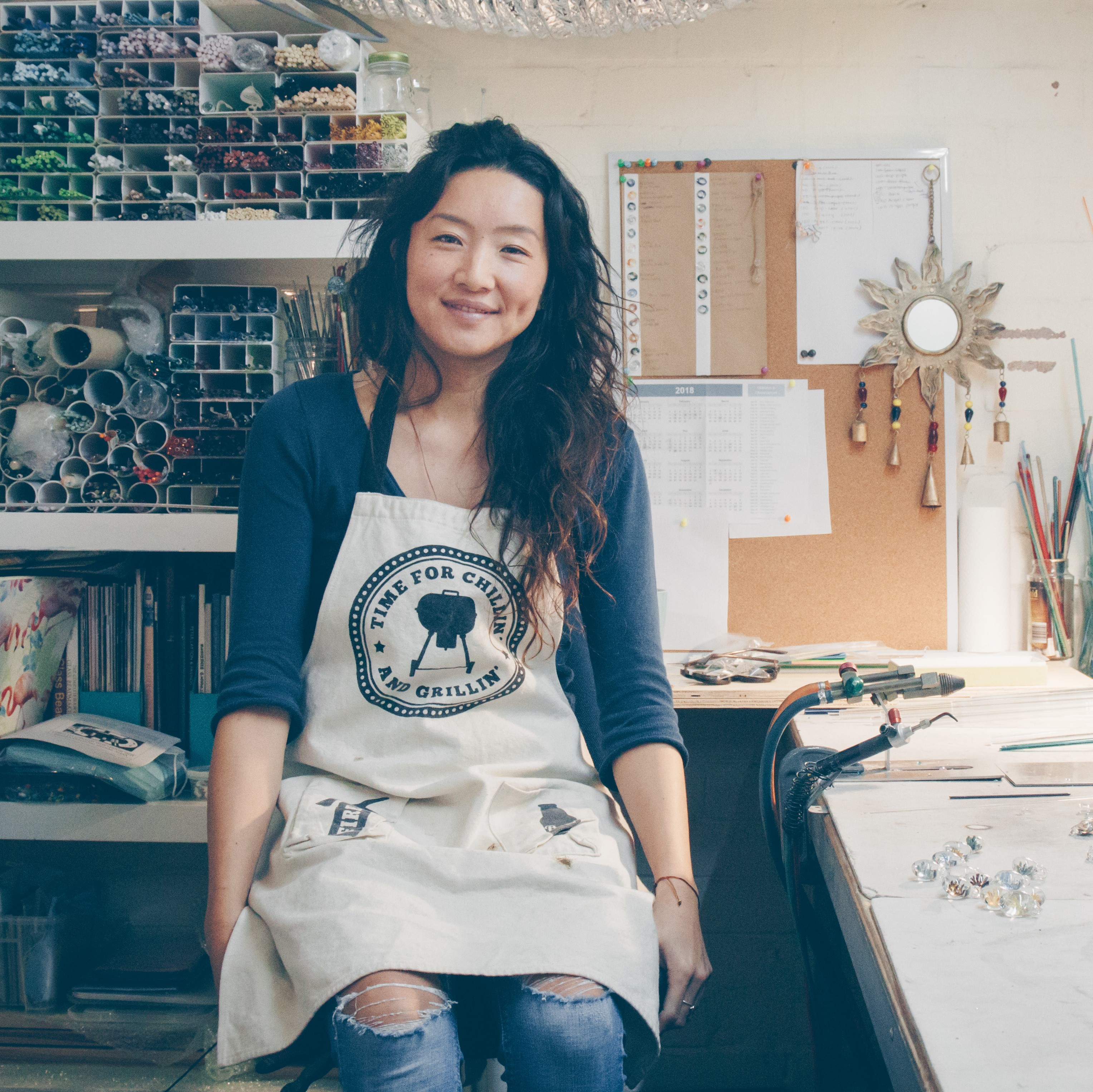 Portrait photo of Mai, sitting on a stool with colourful glass rods in a shelf on her left and glass beads on a desk to her right