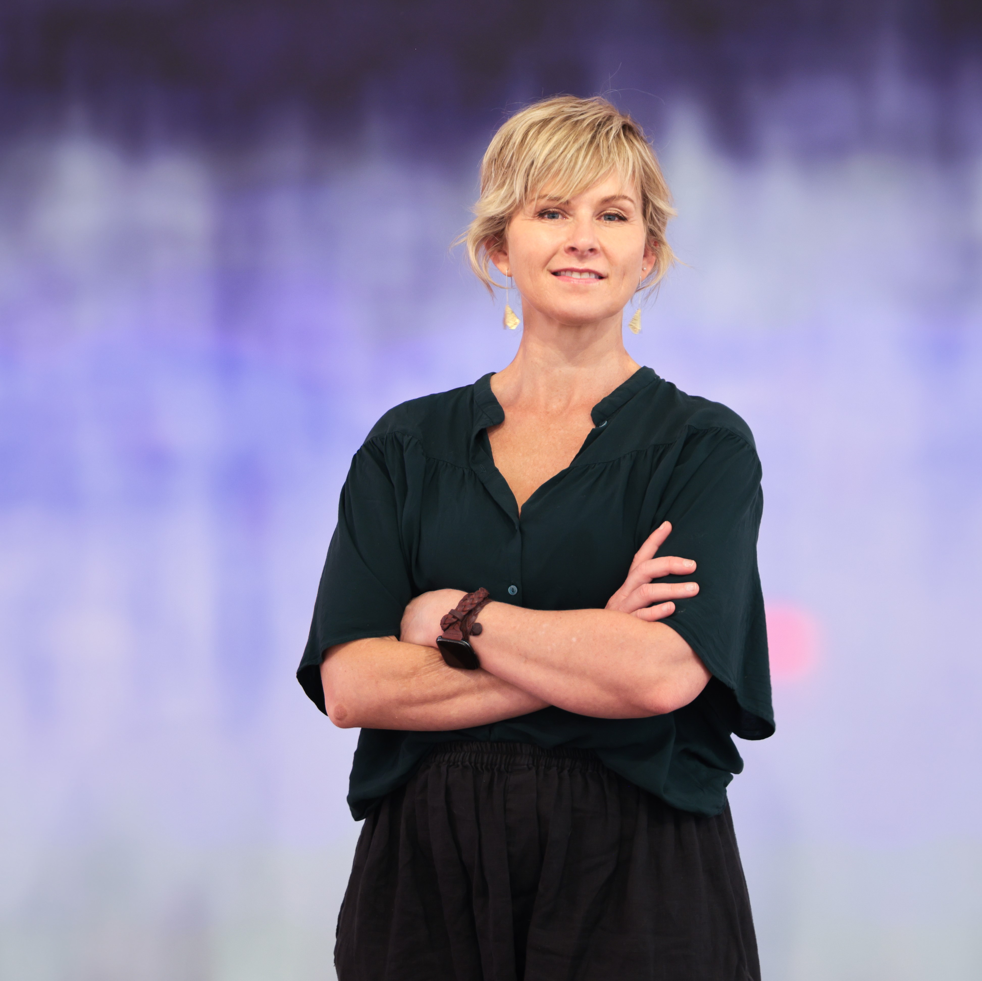 Artist Susie Dureau standing in front of a painting with vibrant purples in an abstract wash, her arms folded and smiling. 