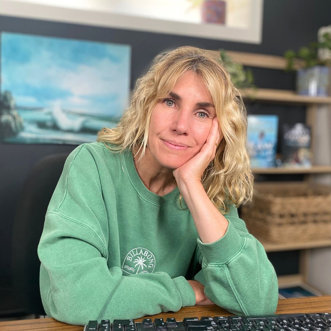 Portrait photo of Kirsty Eager, a woman with wavy blond hair past the shoulders, in a green jumper, sitting down at a desk and looking relaxed and directly at the camera, resting her head on one hand.