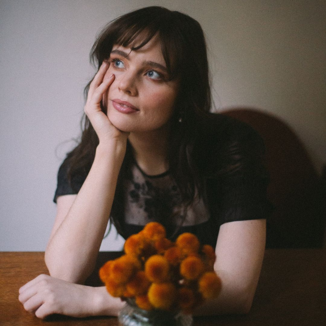 Photo of Kristina Wilson, a young woman with long brown hair, sitting at a table and resting her chin on her hand. A bunch of flowers, possibly billy-buttons, sit in the foreground.