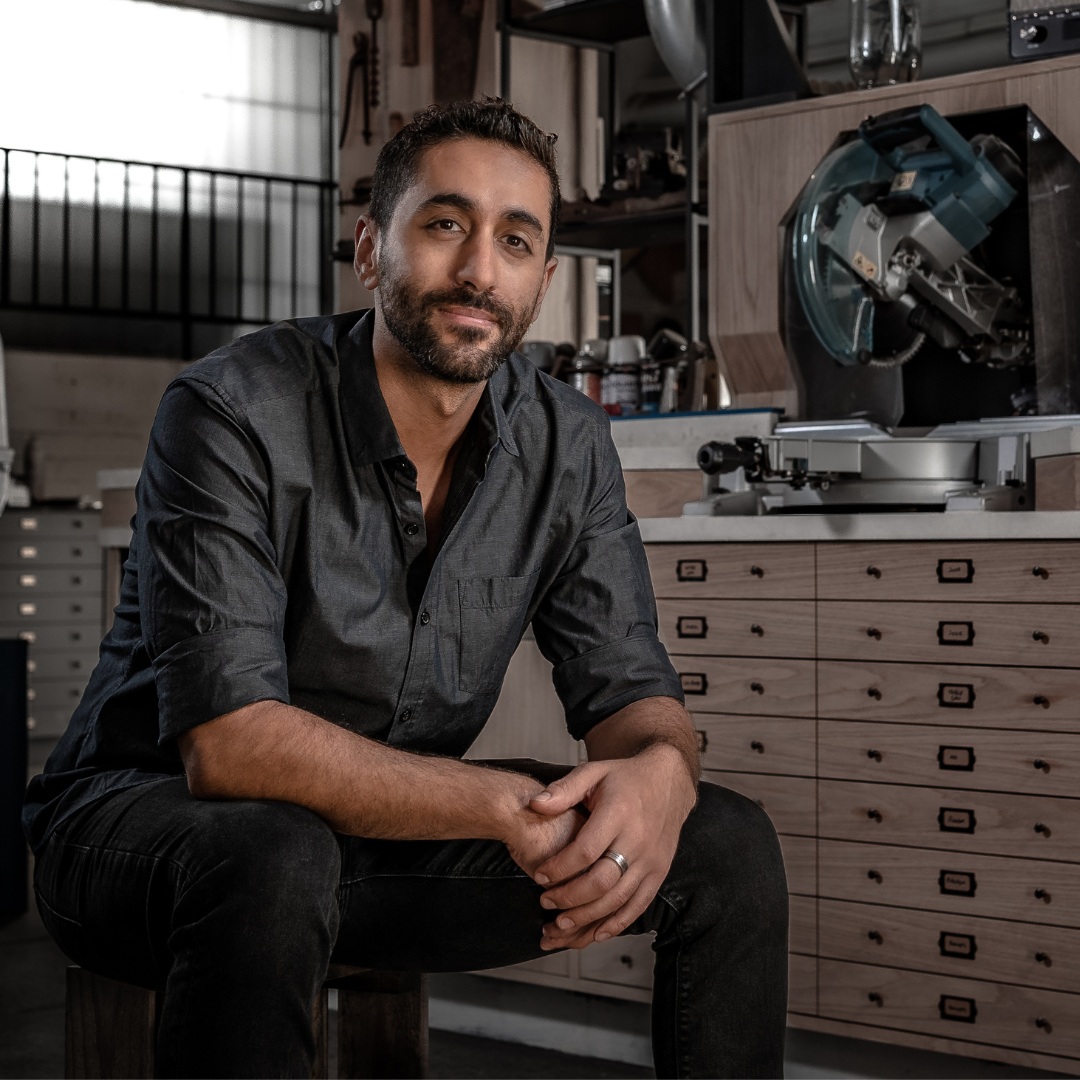 Nick casually sits in a tidy timber workshop, surrounded by well-loved and clean power tools and ordered scraps of timber