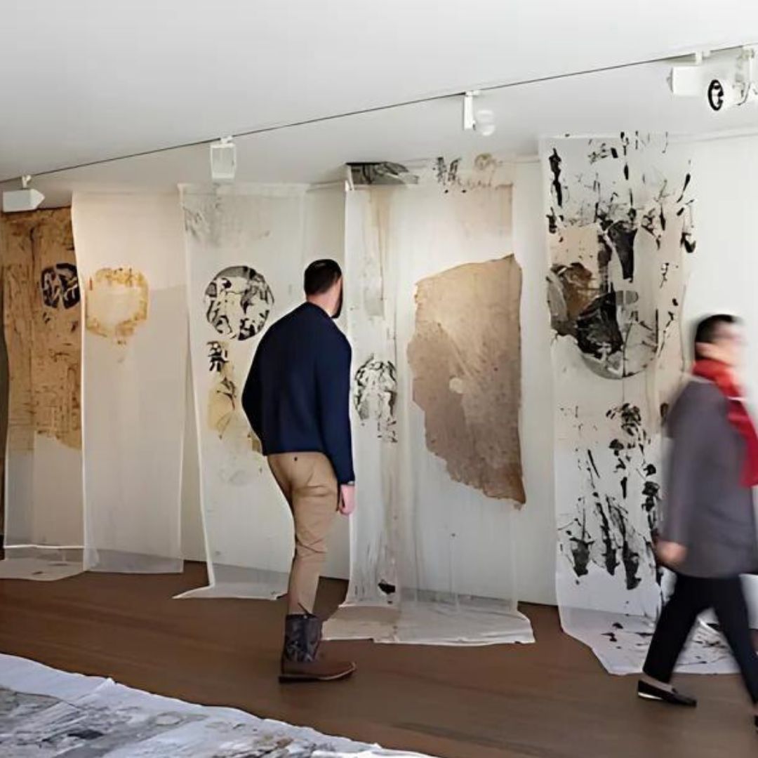 Person in an art gallery looking at fabrics hanging from the ceiling with organic shapes painted on them. Another person is blurred walking out of shot. 