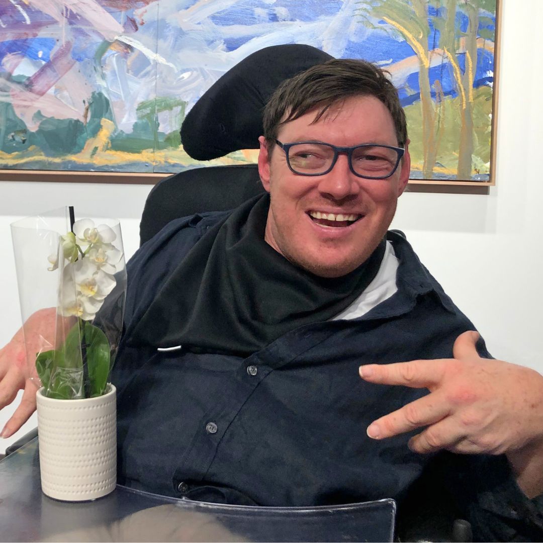 Matthew Elliott smiling at the camera, his artwork in the background.