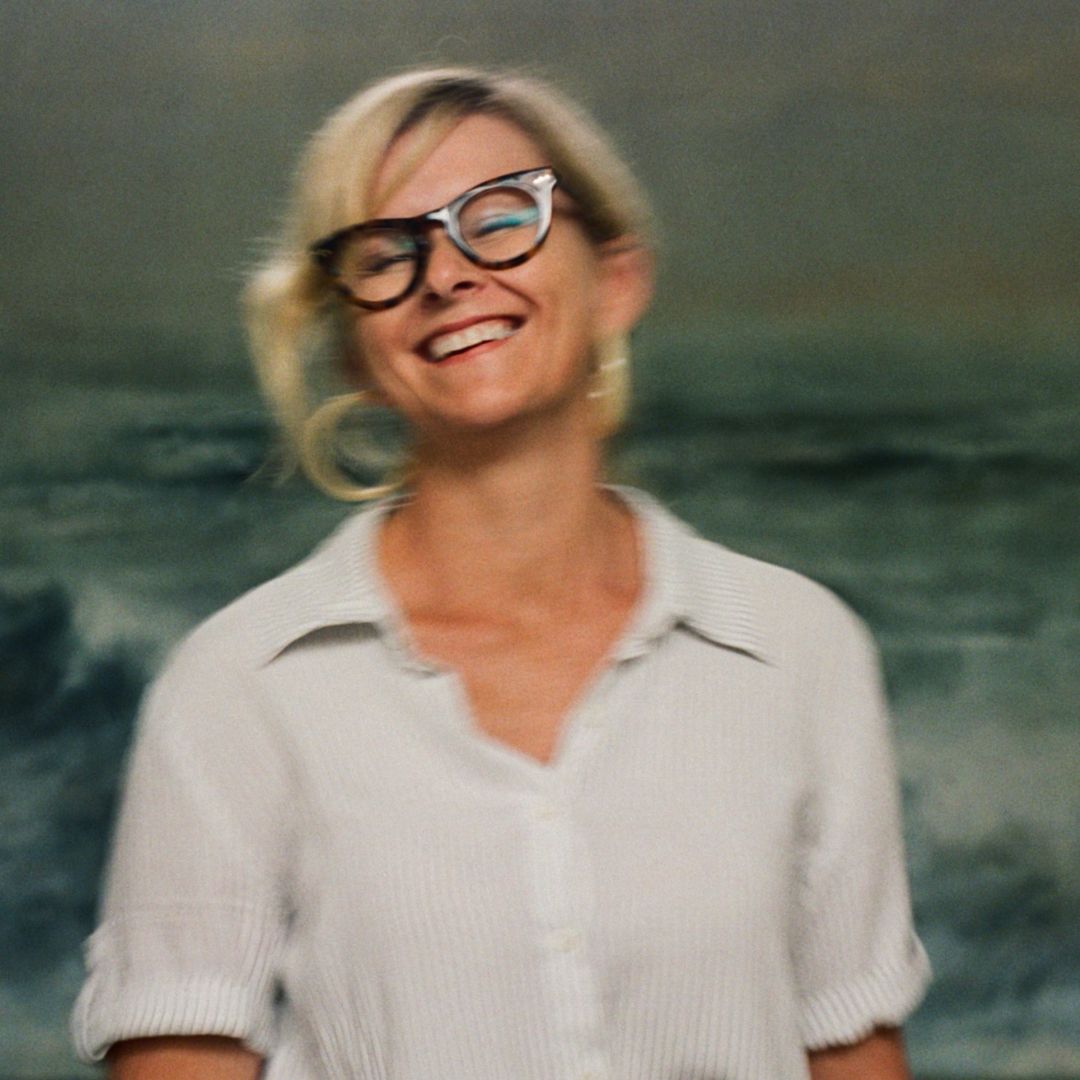 Portrait of Susie Dureau smiling to the camera, wearing thick-rimmed glasses, in front of one of her paintings of a wild ocean