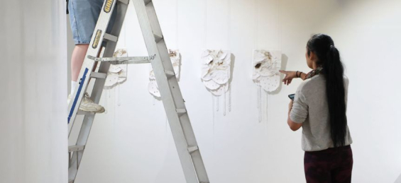 Photo of a person pointing at artworks hanging on a white wall, while another person is standing safely on a ladder and adjusting the artworks. 