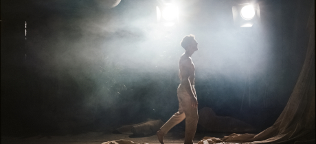 Male dancer walking through a misty space, theatre lights silhouetting him, heavy canvas laying on the ground around him. 