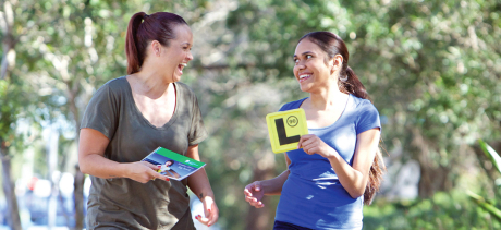  two females looking at each other - one is holding the L plate and the other one learner driver log book