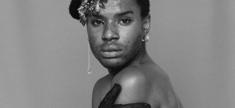 Black and white photo of a person looking directly at the camera, with a strong and focused look in their eyes. Their bare shoulder is directed to camera, they are wearing a beaded headpiece and satin gloves, a long earring on their right ear, and a solid hoop earring on their left.
