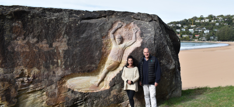 A man and a woman stand in front of a stone carving of a man surfing.