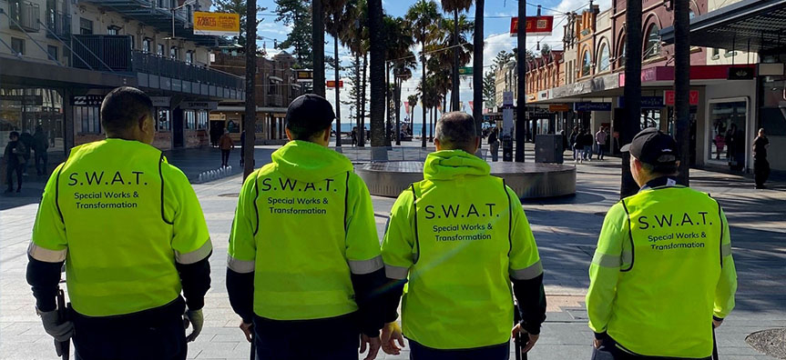 Four Council workers stand on Manly Corso with their backs to the camera wearing high vis vests with S.W.A.T Special Works And Transformation printed on the back