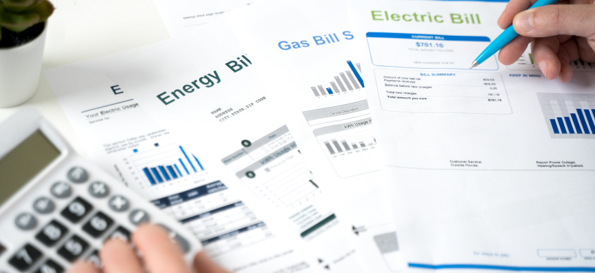 A pile of energy bills on a desk with two hands - one using a calculator and the other holding a pen.