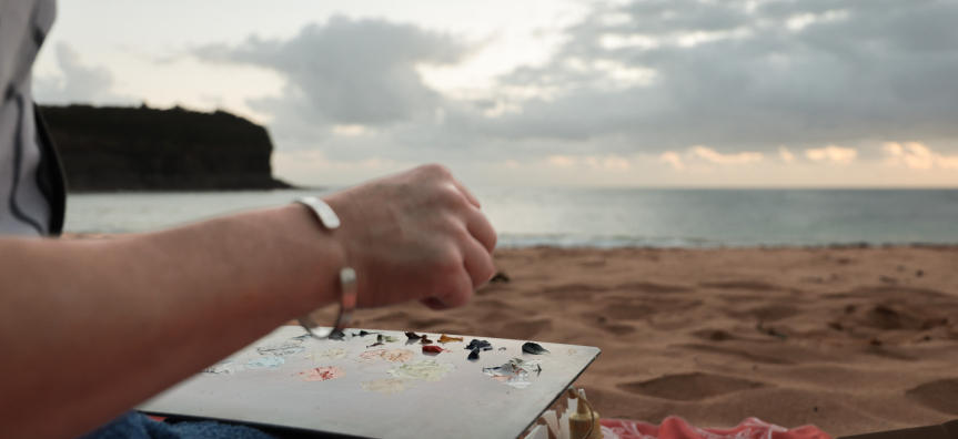 Close up of a hand with a paint palette in the foreground, a beach with the sun rising through it in the background.