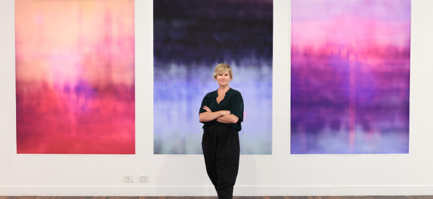 Artist Susie Dureau standing in front of three vibrant abstract paintings, her arms folded and smiling. 
