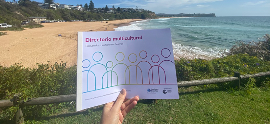 A print version of the multicultural directory is held by a hand with Warriewood Beach in the backgroun