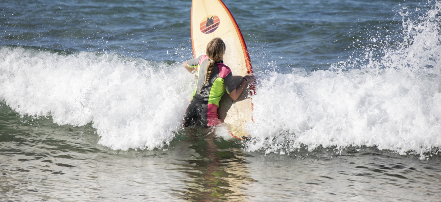A young actor launching into a shore-breaker with her surfboard. 