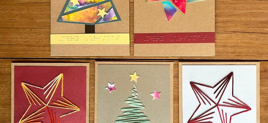 Five Christmas cards on a timber table, each with brightly coloured cut out shapes forming Christmas trees and stars.