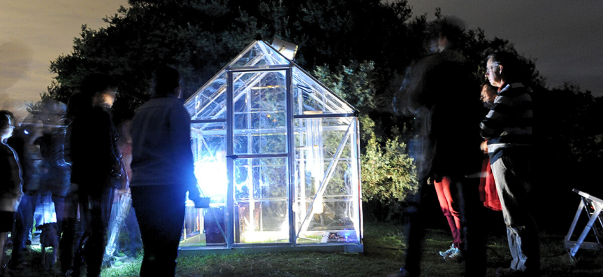 Photo at night of a small-scale glasshouse light up from the inside, with people standing around looking into it. 