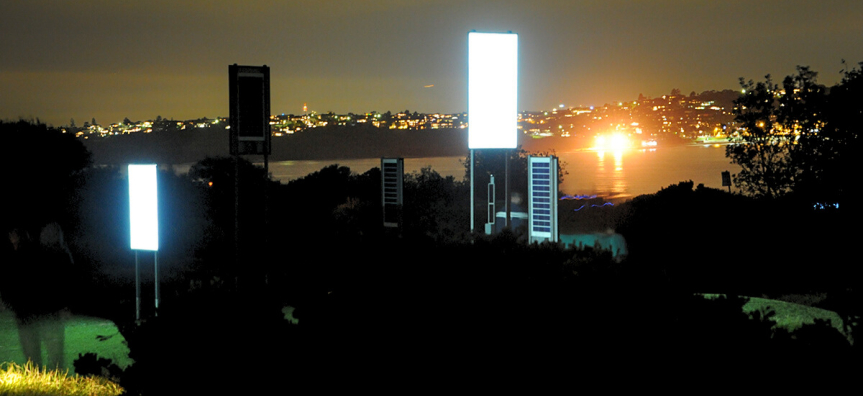 Photo at night, looking over Middle Harbour, large rectangle panels of light brightly shining in the foreground, placed among the vegetation