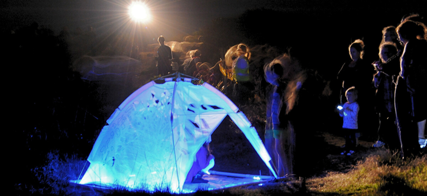 Photo at night, a brightly glowing two-person dome tent sits in the grass, people wandering in and around it. 