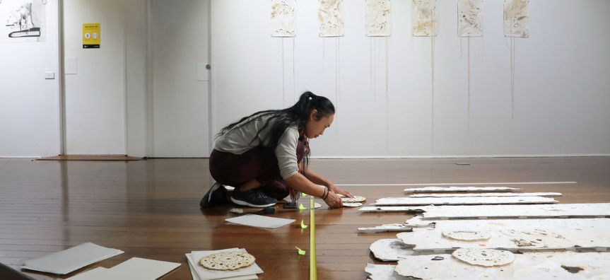 Person kneels on the ground, placing pieces of paper along a measuring tape. Artworks hang evenly in the background.