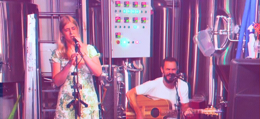 A singer and guitarist perform in front of the machinery of a brewery. 