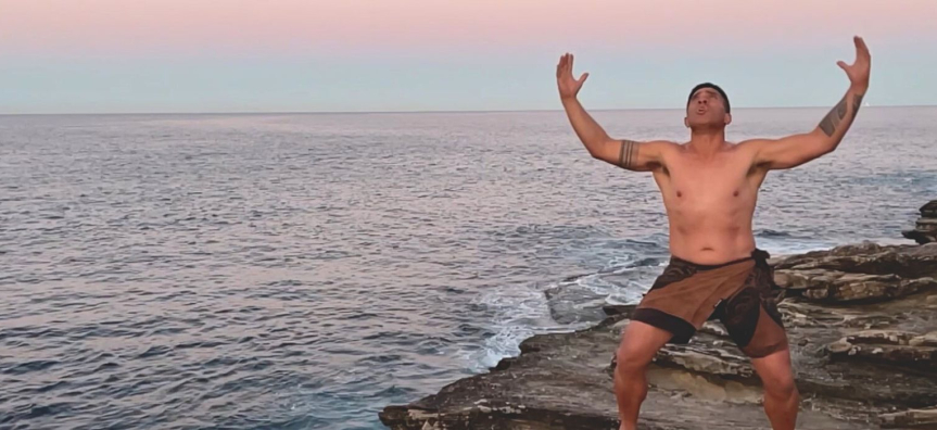 Screenshot from the video artwork Pakoko, showing a male Polynesian dancer performing on the cliffs, arms outreached, next to the ocean at sunrise.