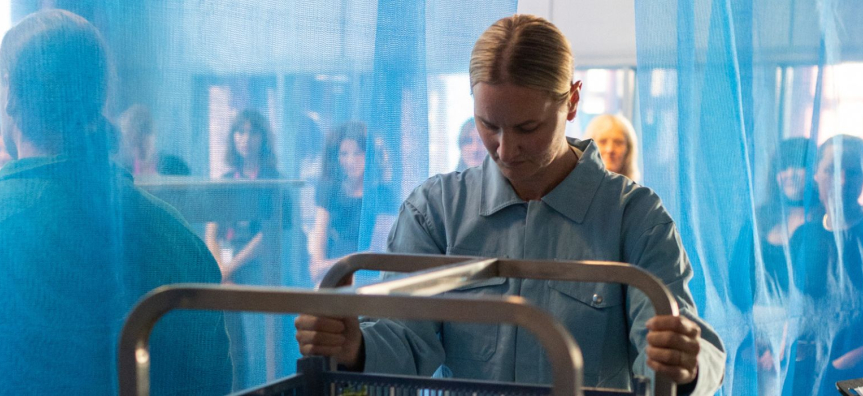 Kate Butcher in the foreground, hair pulled back tightly and wearing blue work uniform, pushing an industrial looking trolley. A sheer blue curtain behind her with a large group of people on the other side, looking like an audience.