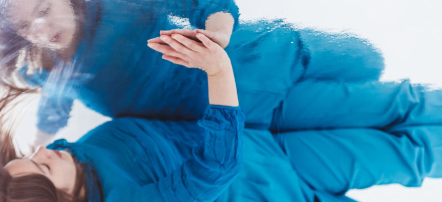 Photo of a dancer in a bright blue tracksuit, pressed against a shiny floor, with a slightly rippled and scuffed reflection.