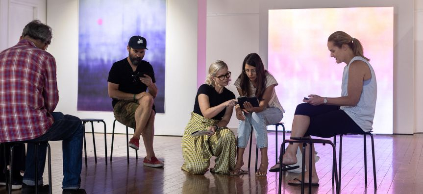 A group of people sitting in an art gallery discussing ideas. Luminescent artworks glow behind them. 
