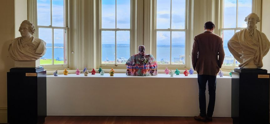 Photo of Daniel Press looking away from the camera, looking at a row of sculptures, which sit in a line in front of a large window.