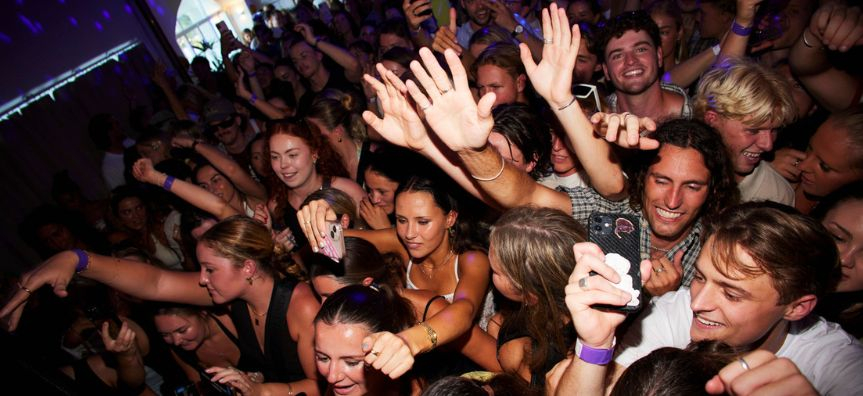 A group of people in the mosh pit at a music performance, hands in the air, mostly smiling.