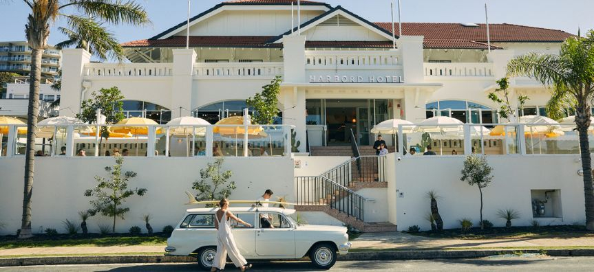 Front of the Harbord Hotel, a large white building with terracotta tiled roof and yellow and white umbrellas across the front. A retro car is parked out front with a longboard on the roof. Two stylish people are walking from the car into the hotel. 