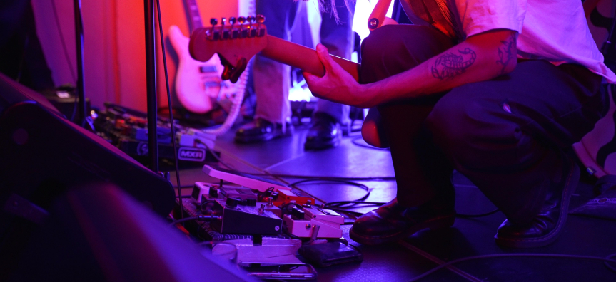 close up of a person kneeling down and playing a guitar under saturated lighting of pink and purple, a scorpion tattoo on their arm