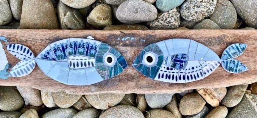 Two mosaic fish made from various patterned tiles facing each other on a piece of weathered timber, sitting on a bed of river pebbles.