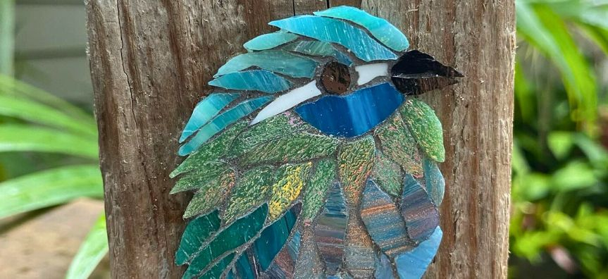 A finely mosaiced blue bird with various reflective tile pieces, shimmering against a piece of weathered timber.