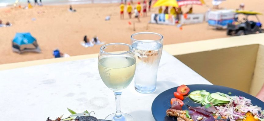 A table with glass of wine, a water, and a plate full of delicious food - overlooking the beach with sand and waves in the background.