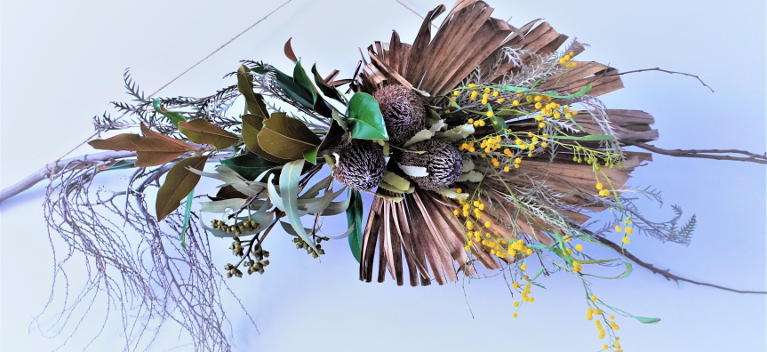 A beautifully composed decorative wall hanging made from dried palm fronds, banksia flowers, and assorted leaves. Fresh bright yellow flowers add a pop of colour. 