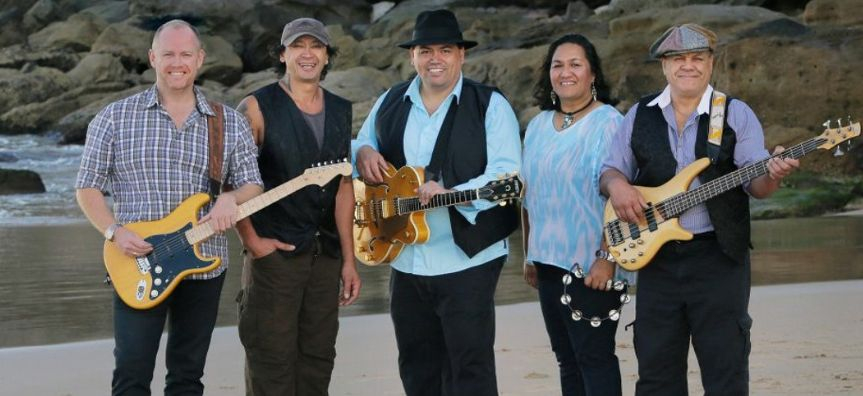 The five band members stand on a beach smiling and holding their instruments. 
