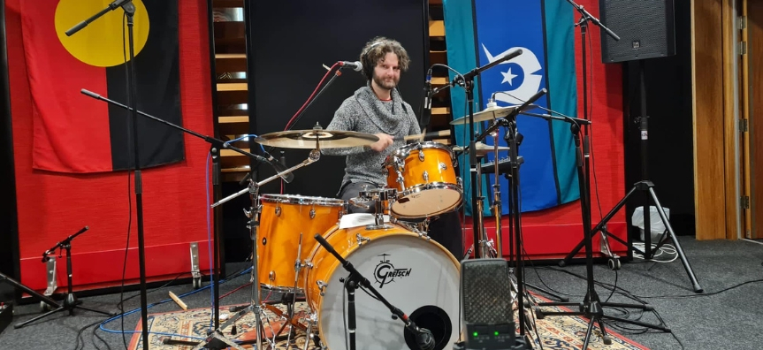 Photo of Patrik Jarlestam in a recording studio playing the drums, with Aboriginal and Torres Strait Island flags draped in the background