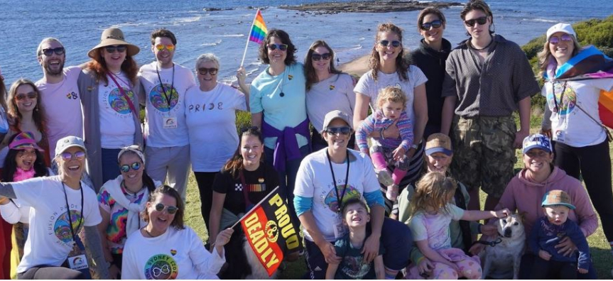Group of brightly dressed smiling people on long reef headland waving rainbow flags