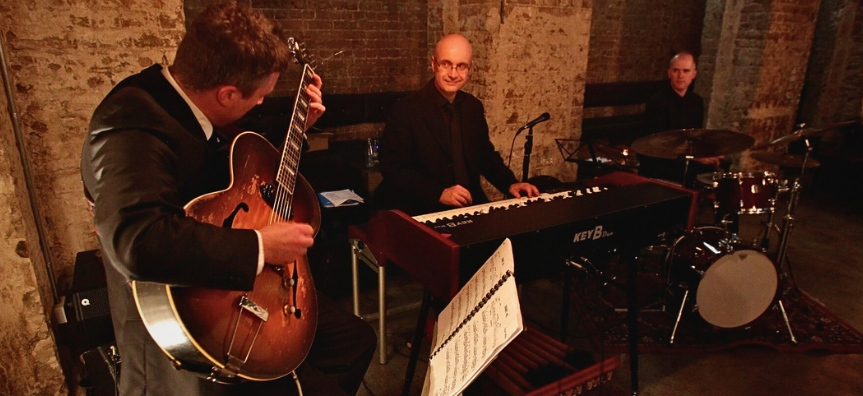 A guitarist, an organist, and a drummer playing against a rustic brick wall, smiling at each other