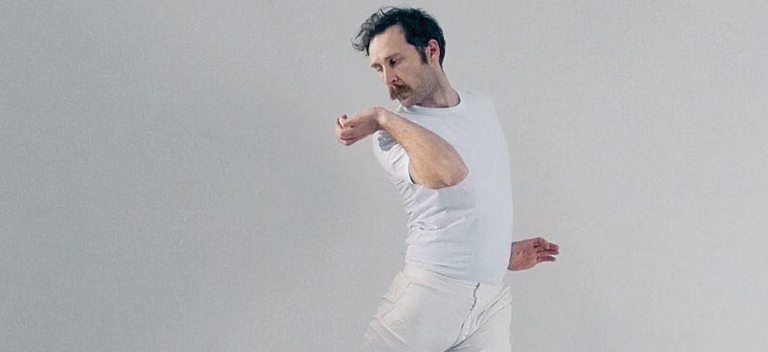 A man is mid-dance, arching his back with front hand stretched backwards to accentuate the arch. He wears a white t-shirt and jeans, against a white background. His moustache stands out.