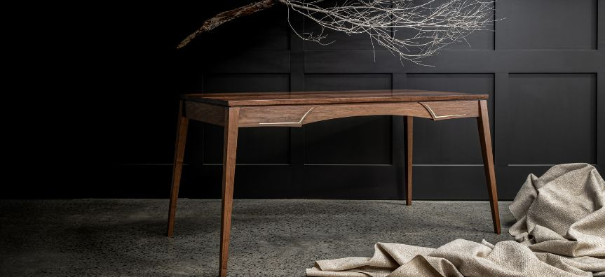 A delicate, almost art nouveau timber writing desk with brass inlay against a black wall, with a large and delicate tree branch hanging overhead