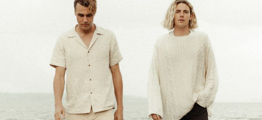 Two people walking down a beach, one in a white short sleeve button up shirt looking down, the other with a white knitted jumper looking to the camera
