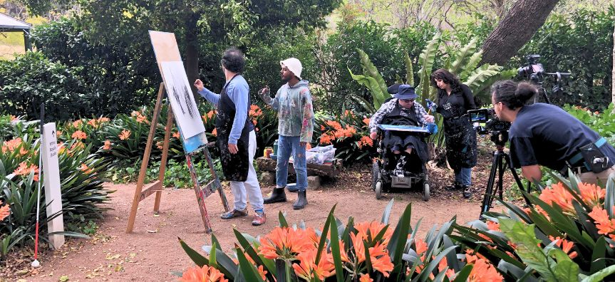 Matthew Elliott with collaborators painting outside, surrounded by a lush garden.