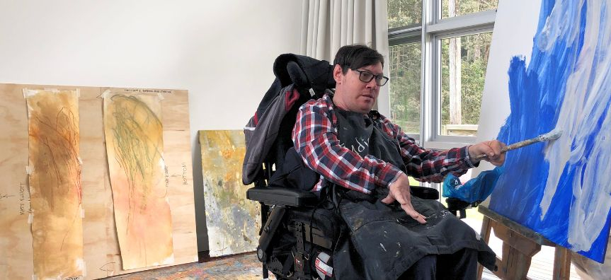 Artist Matthew Elliott, sitting in a wheelchair, painting a large canvas with broad brush strokes, smaller artworks leaning on the wall behind him