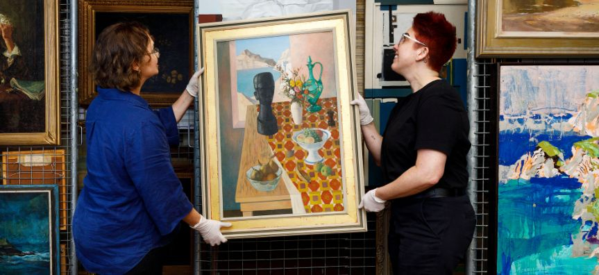 Two people stand either side of a painting, lifting it onto a rack. They are wearing white gloves and surrounded by other paintings.