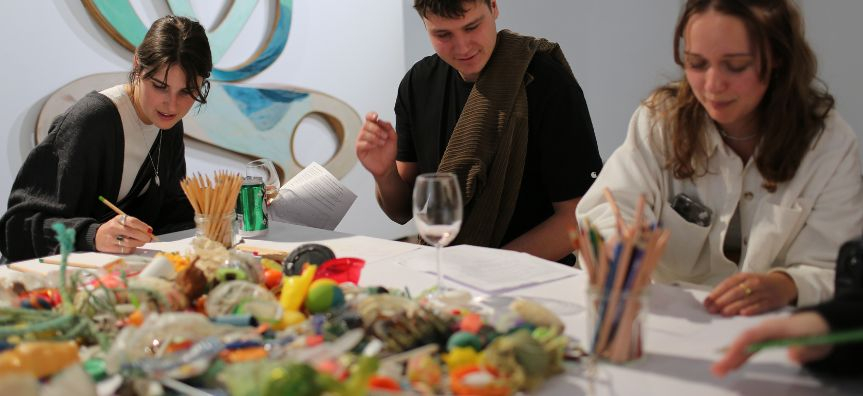 three people sit at a table drawing found plastic objects on the table