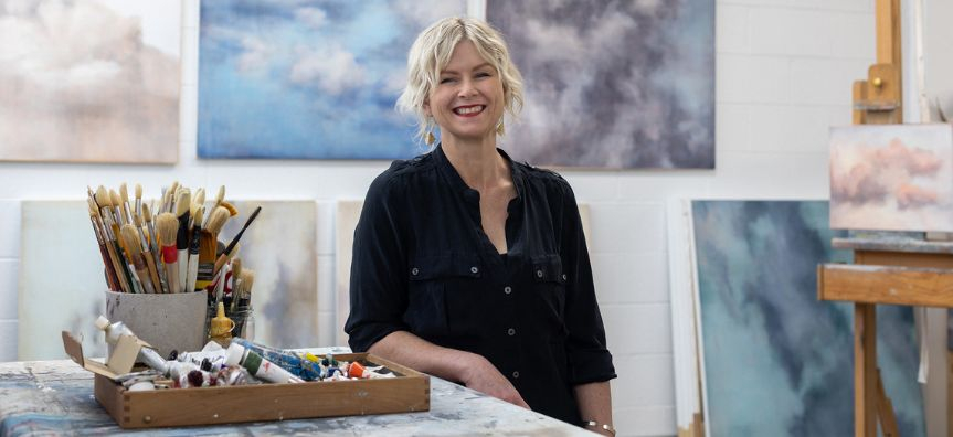 Susie stands in a studio surrounded by paintings of cloudy skies, paints and brushes sitting on the table next to her. 