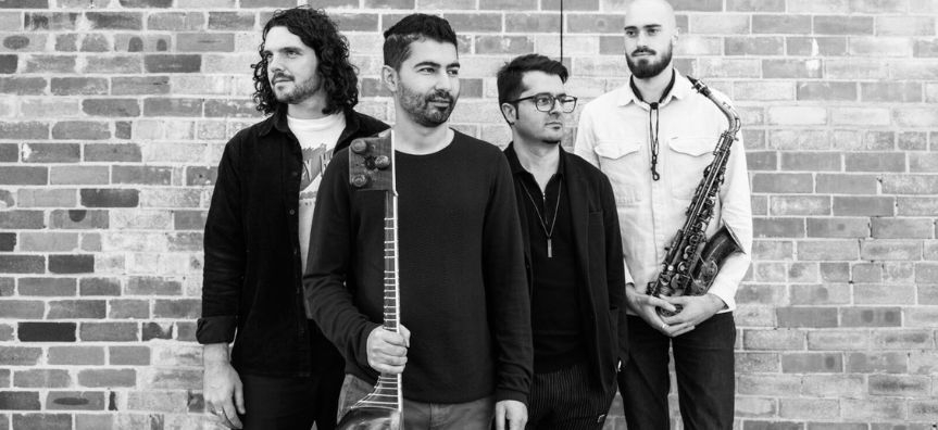 four musicians stand in front of brick wall looking in different directions, two holding instruments